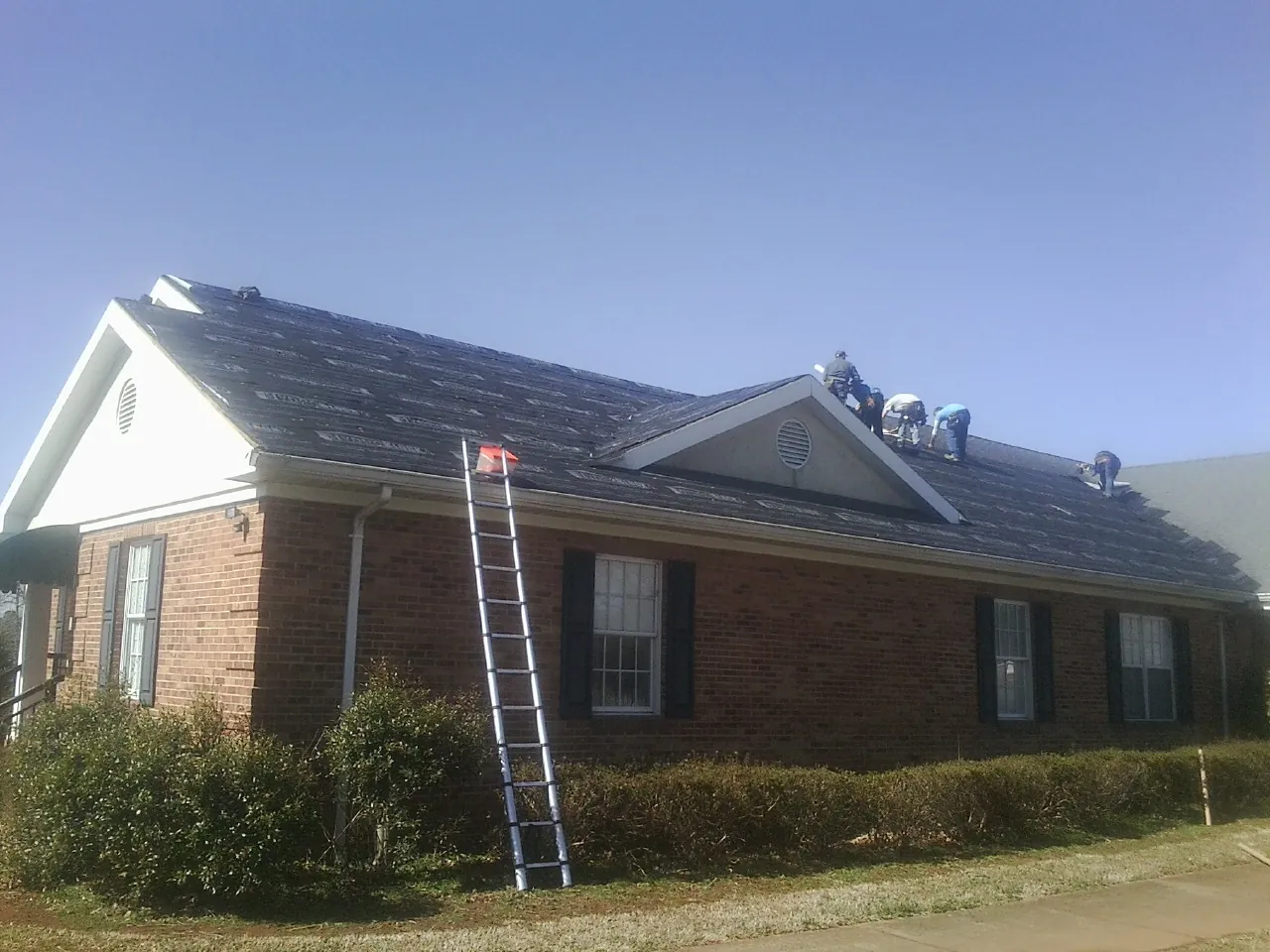 Crew tearing shingles off a brick ranch roof with a ladder set — Emergency Roof Repair work by our crew in Hedrick