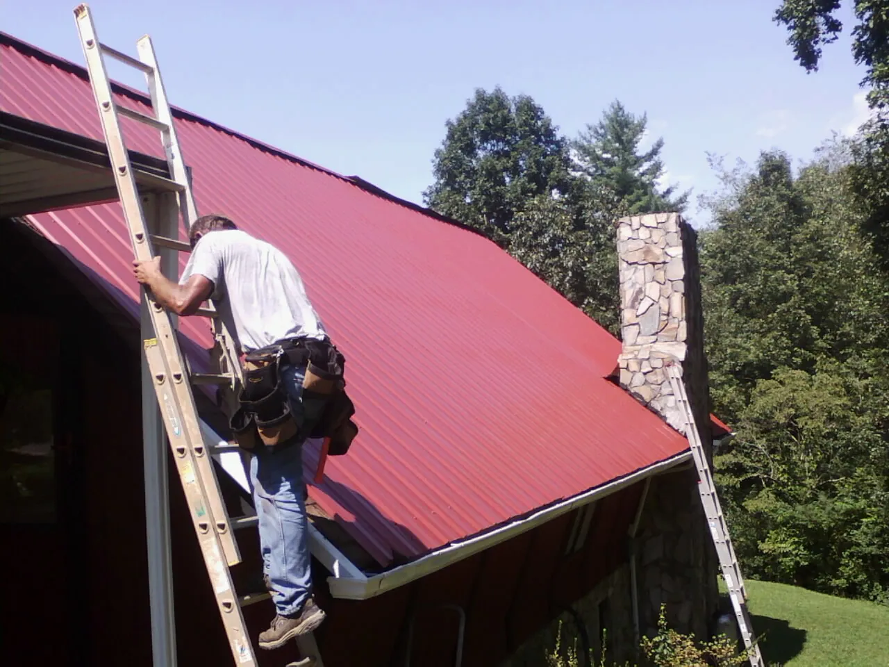 Roofer on a ladder beside a red metal roof and stone chimney — Soffit & Fascia Repair by Roofing Brothers in Hedrick